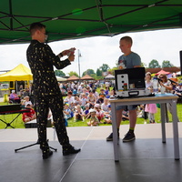 Na zdjęciu stojący na scenie iluzjonista wraz z chłopcem bioracym udział w sztuczce,  w tle widoczna publiczność
