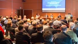 Fotografia przedstawia salę konferencyjną ze słuchaczami siedzącymi na krzesłach, skierowanymi w stronę ekranu z prezentacją.